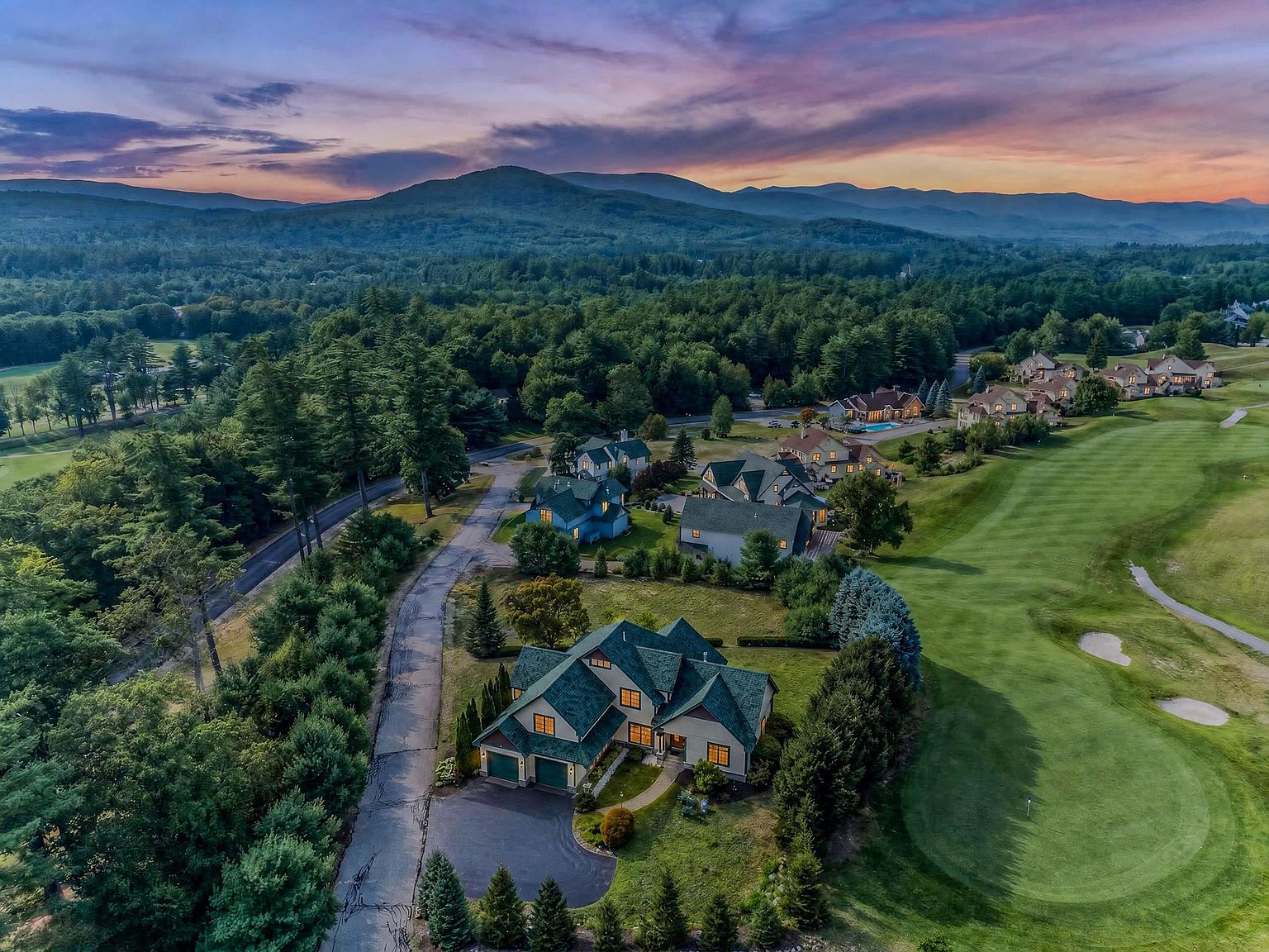 This stunning aerial perspective captures a sprawling residential property nestled alongside a lush, manicured golf course. The home features a multi-gabled roofline with dark green shingles and a classic exterior, surrounded by dense forest and rolling mountain vistas under a dramatic sunset sky. The scene emphasizes the peaceful, scenic location and the expansive layout of the neighborhood.