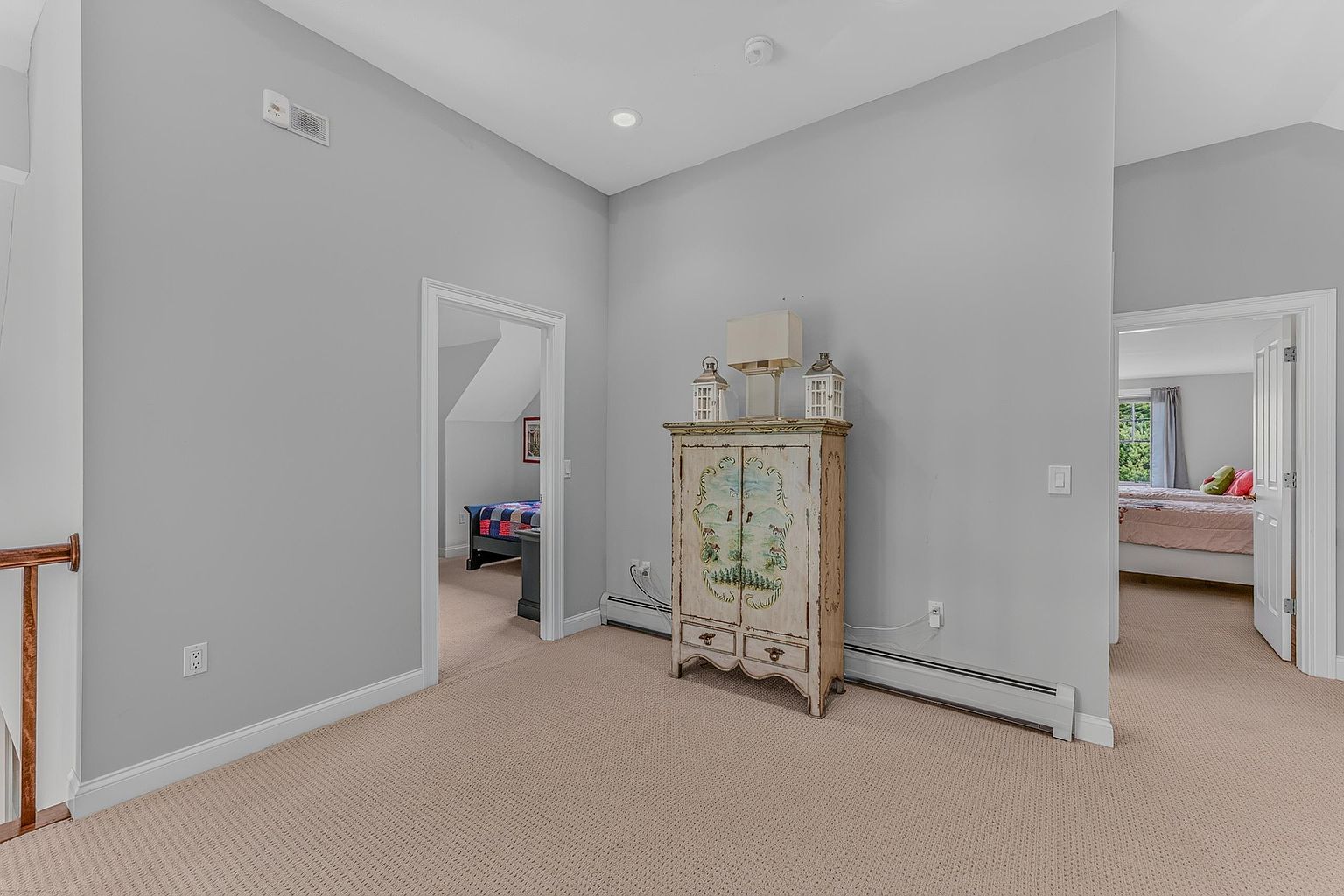 This image shows a carpeted landing area that serves as a central hub connecting two separate bedrooms. A decorative, hand-painted wooden cabinet stands against the central wall, topped with a lamp and two lanterns, adding a touch of character to the neutral gray space. The perspective is from the top of a staircase, looking across the landing toward the open doorways of the adjacent rooms.