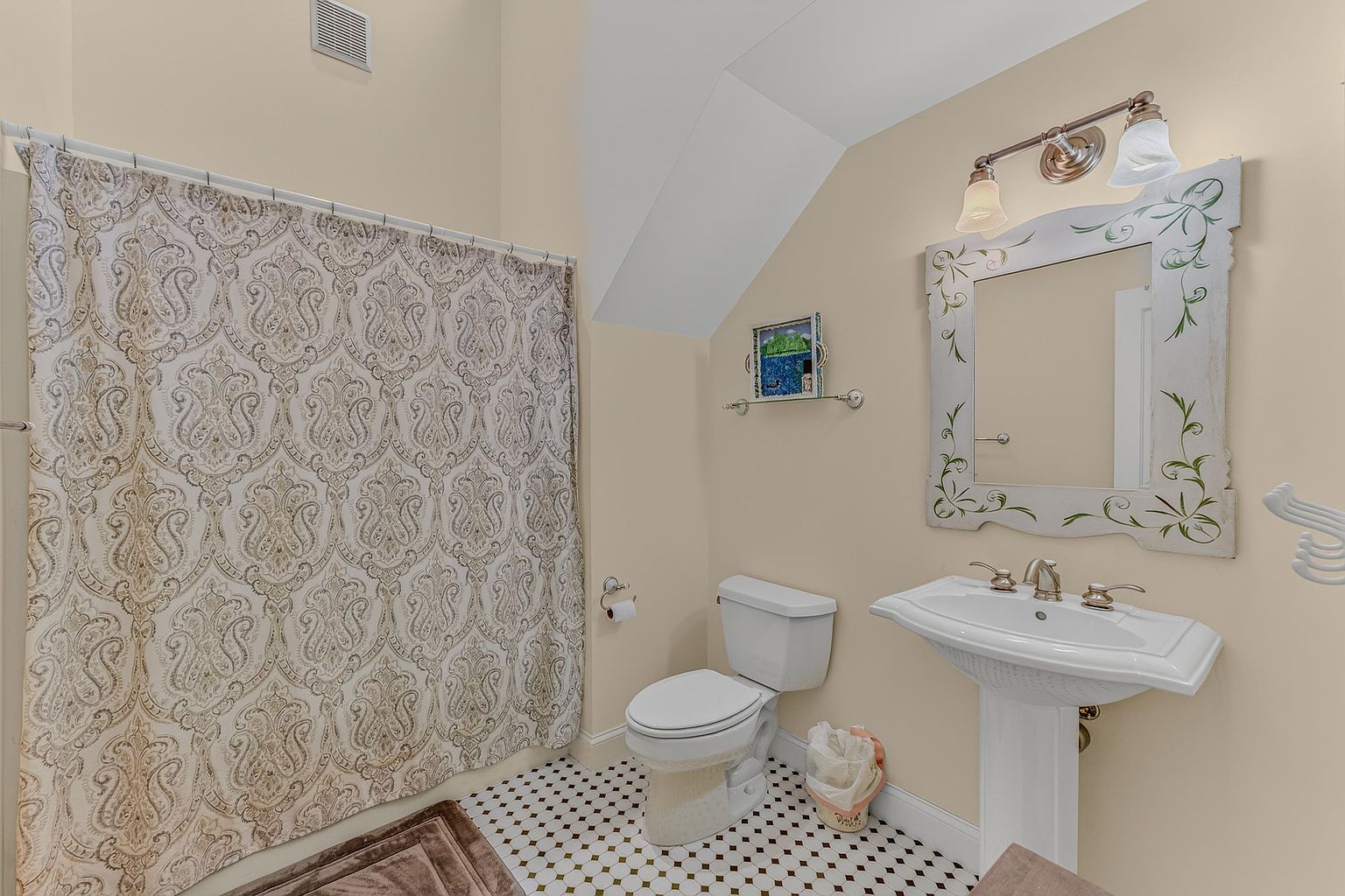 This compact bathroom features a pedestal sink with a decorative mirror and a shower area enclosed by a patterned shower curtain. The space is finished with a classic black-and-white diamond-patterned tile floor and light-toned walls. The room is illuminated by a multi-bulb vanity light fixture above the mirror.