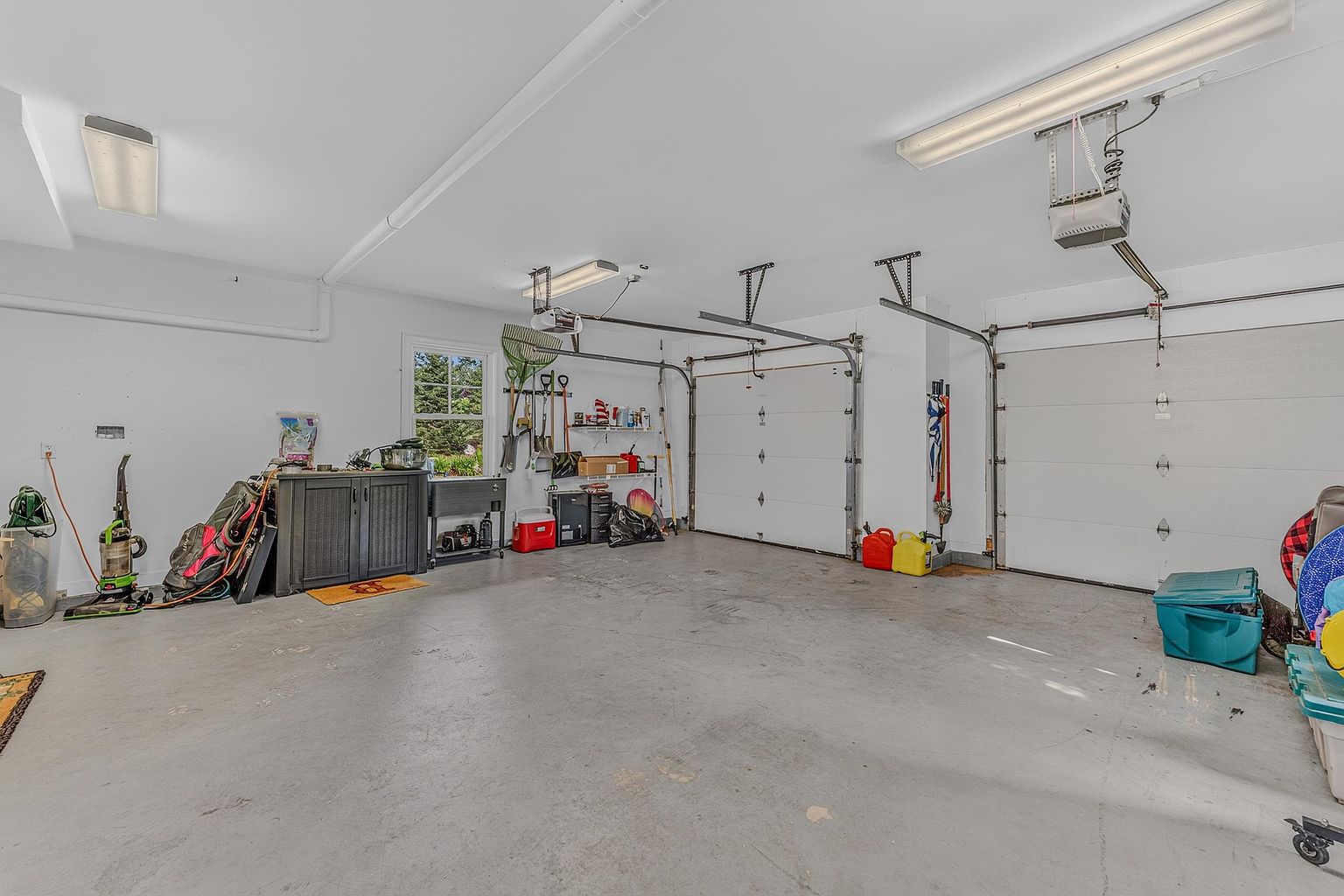 This spacious two-car garage features a clean, neutral aesthetic with light-colored walls and a smooth concrete floor. The space is equipped with two overhead garage doors, overhead lighting, and ample room for storage, including a dark cabinet unit and various wall-mounted tools. A window on the left wall provides natural light, creating a functional and well-maintained area for vehicle storage or workshop activities.