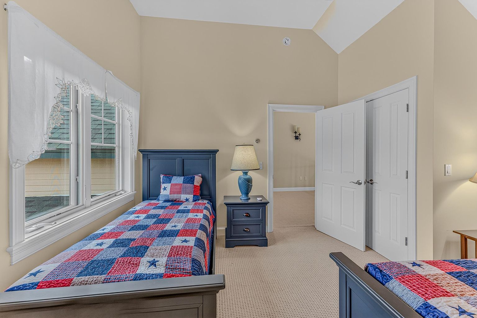 This bedroom features a patriotic theme with two twin beds adorned in red, white, and blue star-patterned quilts. A dark-toned nightstand with a matching lamp sits between the beds, while a doorway leads to an adjacent room. The space is finished with neutral-toned walls, beige carpeting, and a window dressed with a white valance.