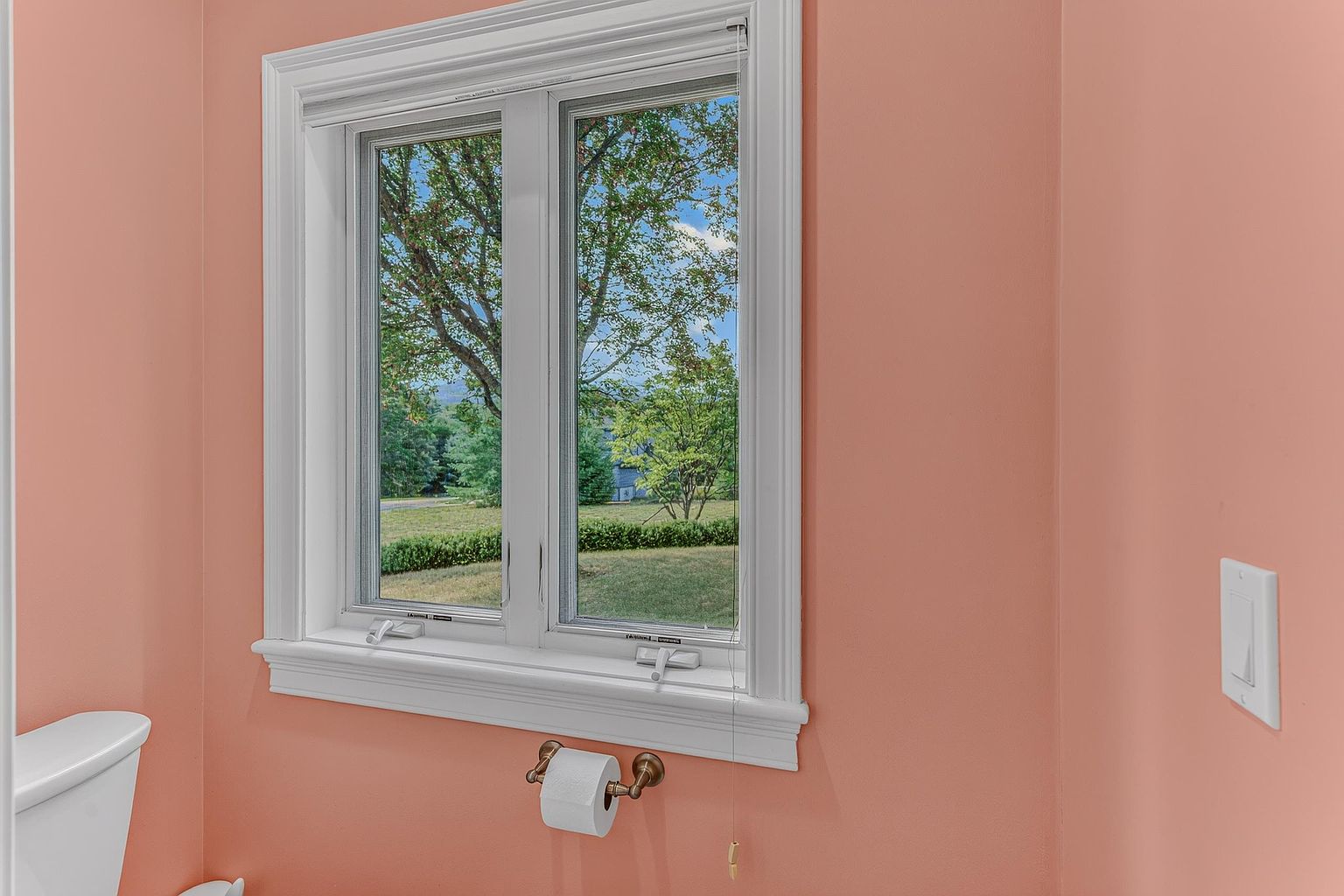 This interior view captures a small bathroom space painted in a soft peach tone, featuring a white-trimmed window that looks out onto a lush green yard. A white toilet is partially visible on the left, and a wall-mounted toilet paper holder is centered below the window. The perspective is eye-level and straightforward, highlighting the simple, functional layout of the room.