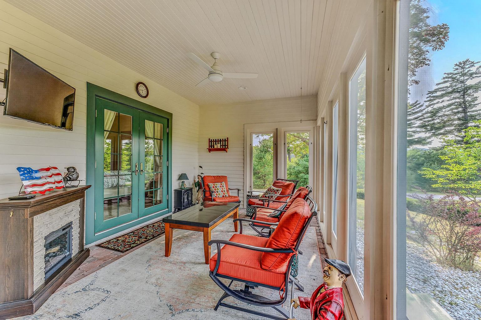 This bright and inviting sunroom features a wall of large windows that provide expansive views of the surrounding greenery. The space is furnished with comfortable orange rocking chairs, a central coffee table, and a fireplace with a stone-clad surround, creating a cozy atmosphere for relaxation. A ceiling fan hangs from the white paneled ceiling, and the room is accented by a decorative figurine and an American flag display.