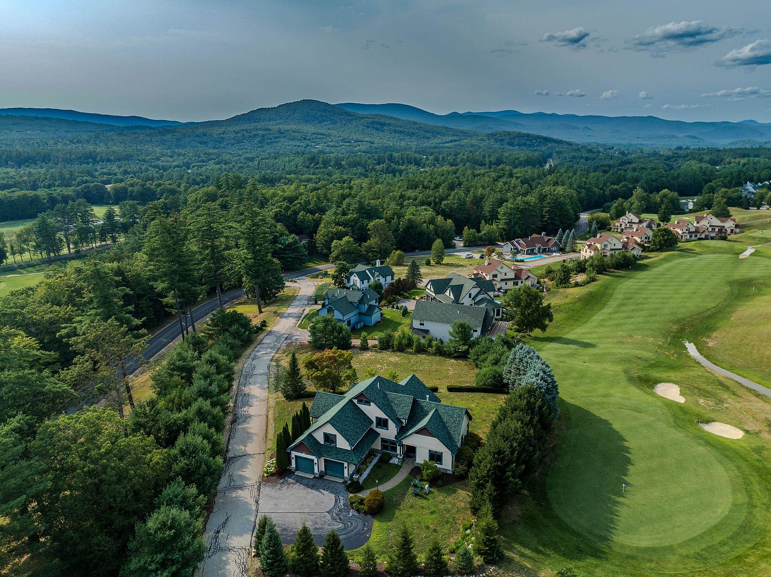This high-angle aerial perspective captures a sprawling residential neighborhood nestled against a lush, forested mountain range. The foreground features a large, multi-gabled home with a dark green roof, situated directly adjacent to a manicured golf course fairway. The scene emphasizes the property's prime location, blending suburban luxury with expansive natural scenery and recreational access.