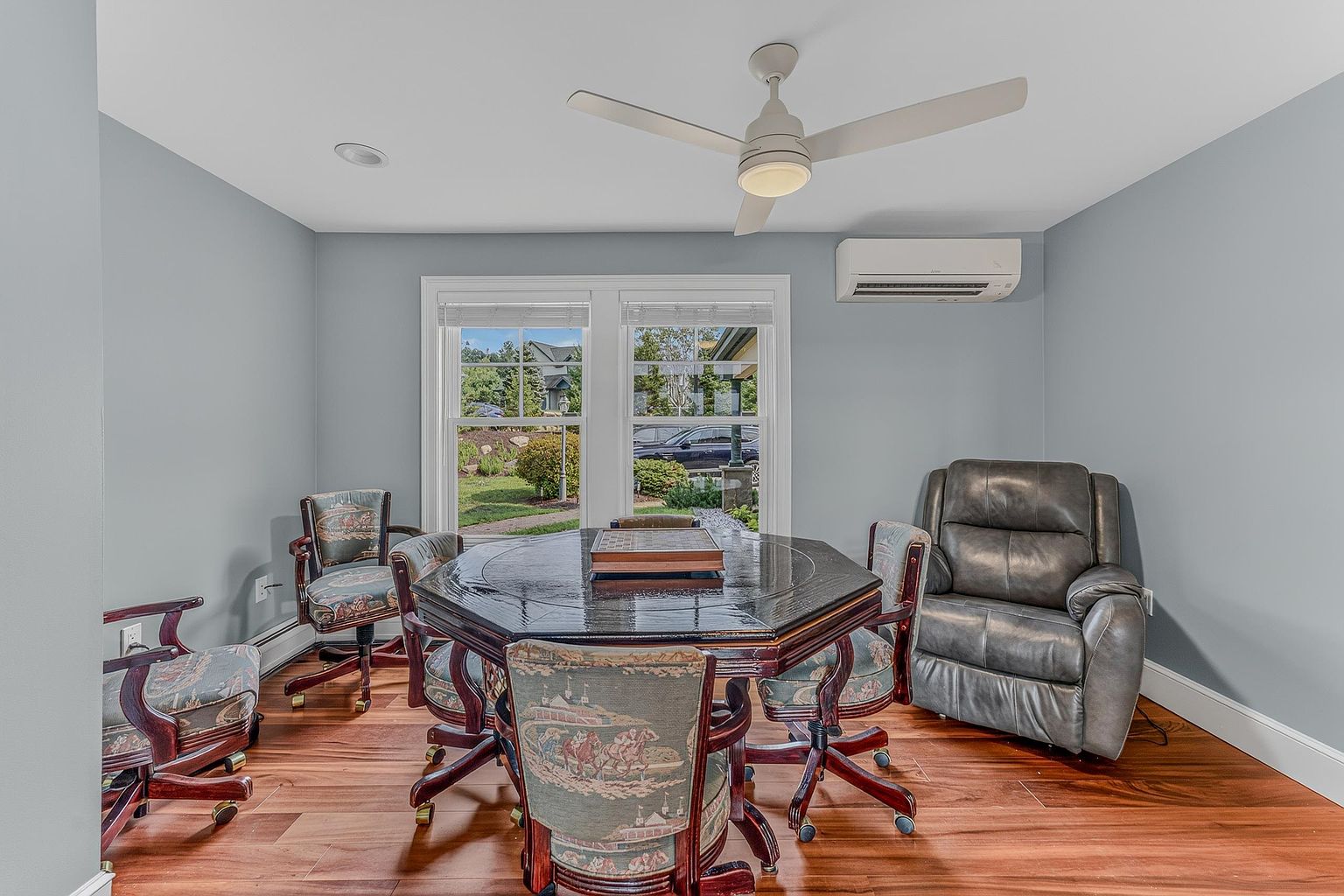 This room functions as a versatile study or game area, centered around an octagonal dark wood table surrounded by four patterned upholstered chairs. A comfortable grey leather recliner sits in the corner, while a ceiling fan and wall-mounted air conditioning unit provide climate control. The space features light blue walls, warm-toned wood flooring, and two windows that offer a view of the outdoor landscape.