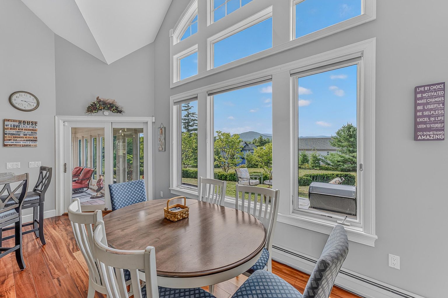 This bright dining area features a round wooden table surrounded by chairs with blue patterned upholstery, set against a backdrop of large windows that offer a scenic view of the outdoors. The room is characterized by its high, vaulted ceiling and light-colored walls, creating an airy and inviting atmosphere. A decorative wall sign and a classic wall clock add a personal touch to the space, which seamlessly connects to an adjacent patio through glass doors.