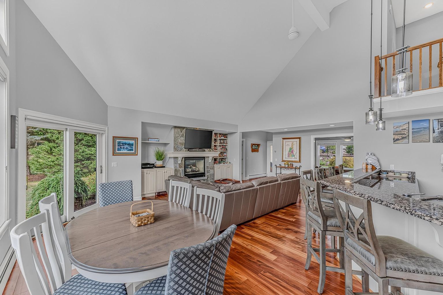 This open-concept living space features a vaulted ceiling and rich hardwood flooring, seamlessly connecting a dining area, a central living room, and a kitchen with a breakfast bar. The living area is anchored by a stone-clad chimney breast with a built-in fireplace and television, flanked by custom cabinetry. The perspective captures the flow of the home, highlighting the bright, airy atmosphere created by the large sliding glass doors and neutral wall tones.