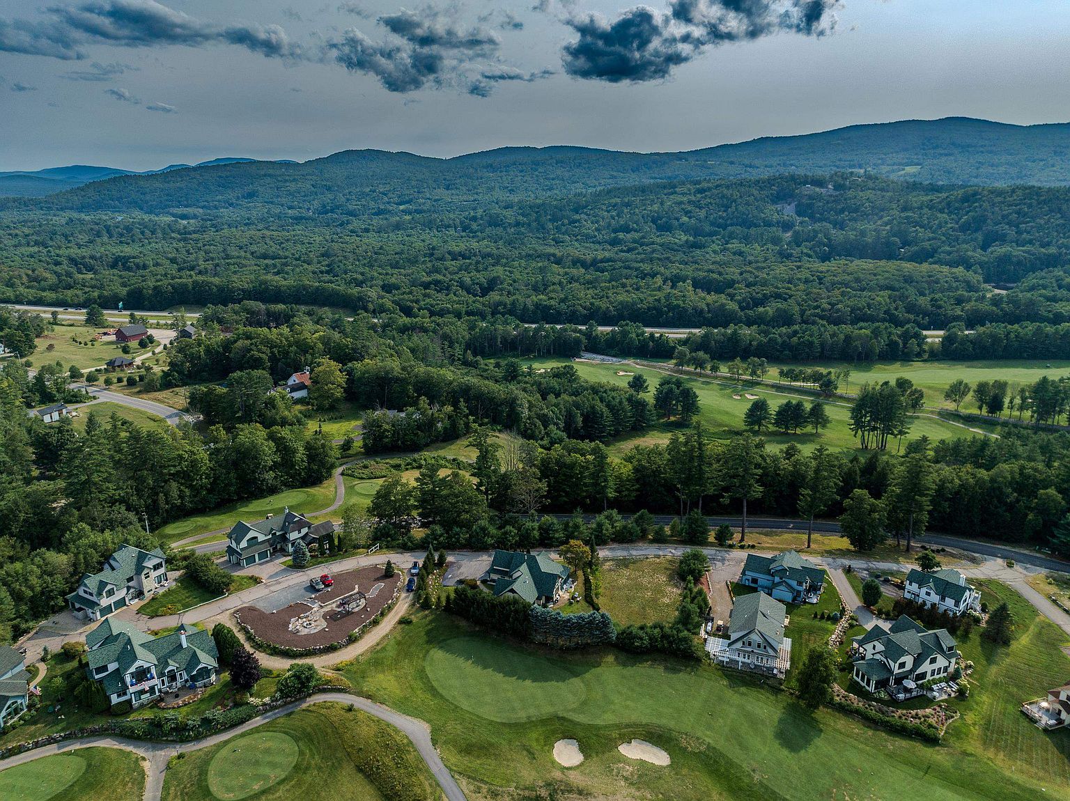 This high-angle aerial view captures a residential community nestled directly alongside a lush, green golf course. The scene features several multi-story homes with dark green roofs, surrounded by mature trees and manicured landscaping, with a vast mountain range visible in the distant background under a dramatic, cloudy sky. The perspective emphasizes the peaceful, scenic integration of the neighborhood with the recreational grounds and natural landscape.