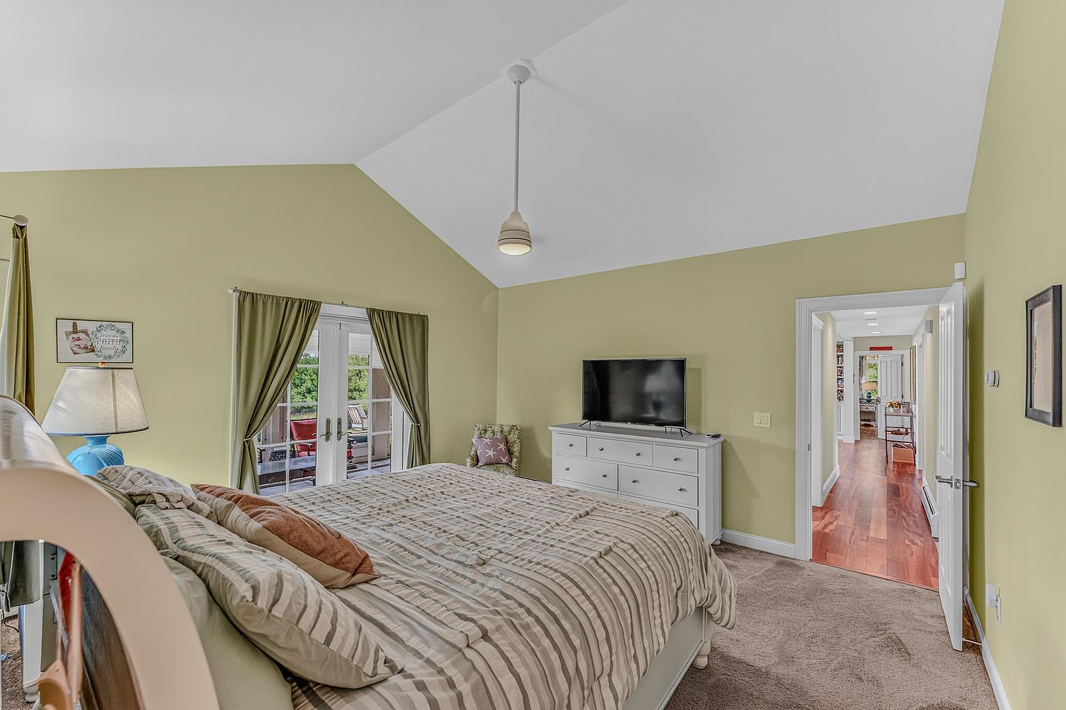 This primary bedroom features a vaulted ceiling and light-colored walls, creating an airy and inviting atmosphere. A large bed with a striped comforter serves as the focal point, positioned near a white dresser topped with a television. French doors with green curtains lead to an outdoor space, while a doorway provides a view into a hallway with hardwood flooring.
