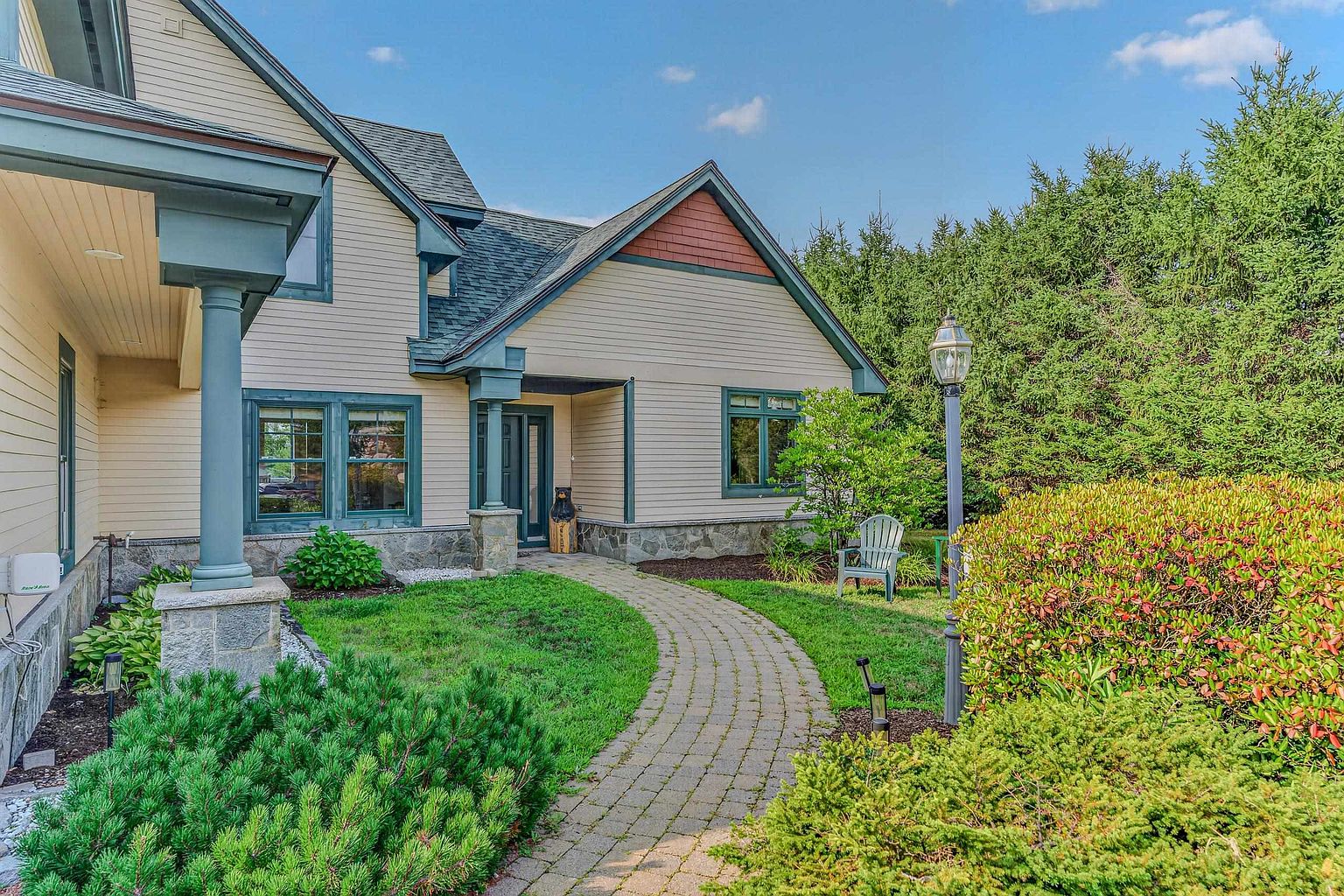 This front exterior view showcases a charming home with light-colored horizontal siding, stone accents at the base, and dark-trimmed windows. A curved stone paver walkway leads to the recessed entryway, which is framed by sturdy columns and accented by a decorative bear statue. The scene is completed by a lush green lawn, mature landscaping, and a tall lamp post, creating a welcoming and well-maintained curb appeal.