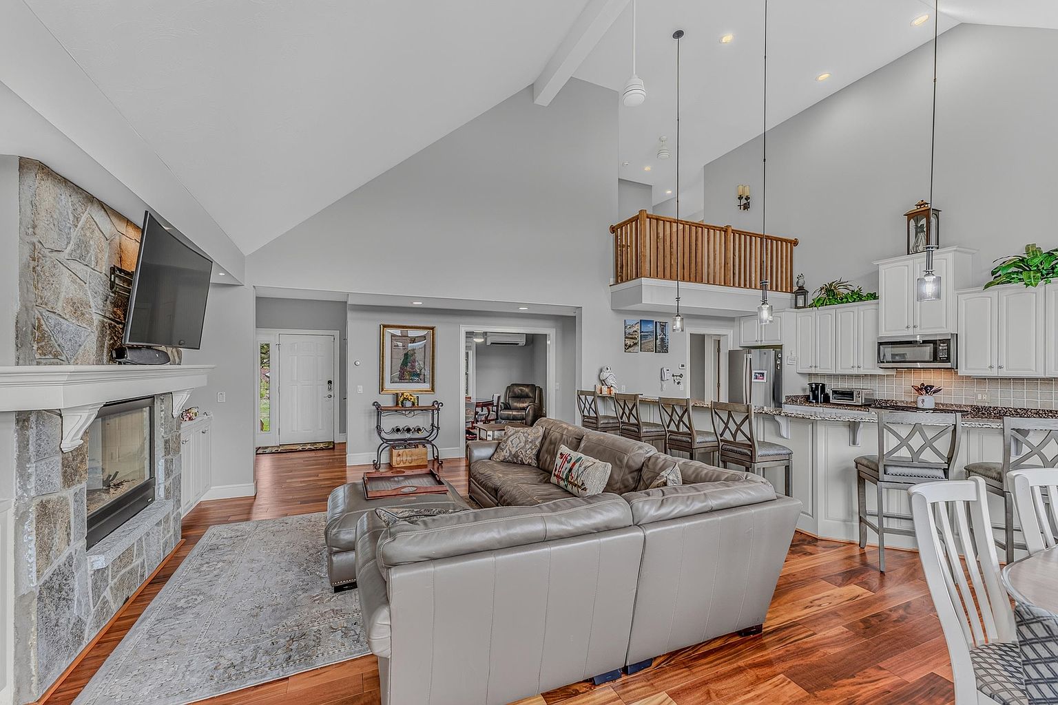 This open-concept living area features a vaulted ceiling and a prominent stone-clad chimney breast housing a fireplace. The space flows seamlessly into a kitchen with a breakfast bar, while a wooden loft railing overlooks the main living area from above. Warm wood flooring and neutral walls create a cohesive, inviting atmosphere suitable for modern family living.