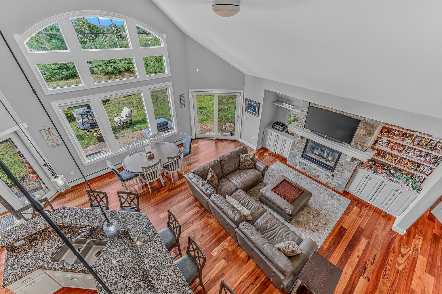 This high-angle shot captures an open-concept living and dining area characterized by vaulted ceilings and rich, warm-toned wood flooring. The space features a large leather sectional sofa arranged around a central ottoman, positioned near a stone-clad chimney breast with an integrated television and fireplace. A dining set sits adjacent to a wall of large windows that provide abundant natural light and views of the outdoor patio, creating an airy and inviting atmosphere.