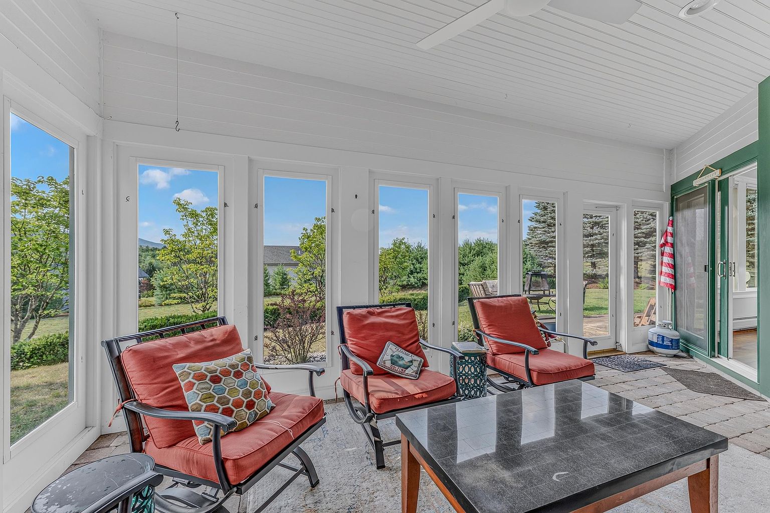 This bright and airy sunroom features a series of large windows that provide expansive views of the surrounding landscape. The space is furnished with three comfortable armchairs with orange cushions, a patterned accent pillow, and a central dark-topped coffee table. The room has a light-colored ceiling with a ceiling fan and a paved floor, creating a relaxed, inviting atmosphere perfect for indoor-outdoor living.