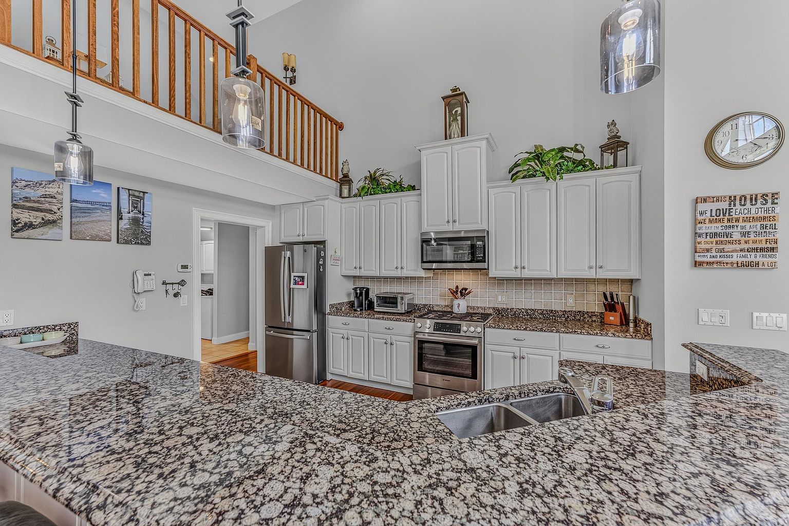 This bright and open kitchen features white cabinetry paired with speckled stone countertops and a stainless steel appliance suite. A large peninsula provides ample workspace and seating, while a lofted area with a wooden railing overlooks the space from above. The room is accented by decorative wall art, a wall-mounted clock, and pendant lighting, creating a warm and inviting atmosphere.