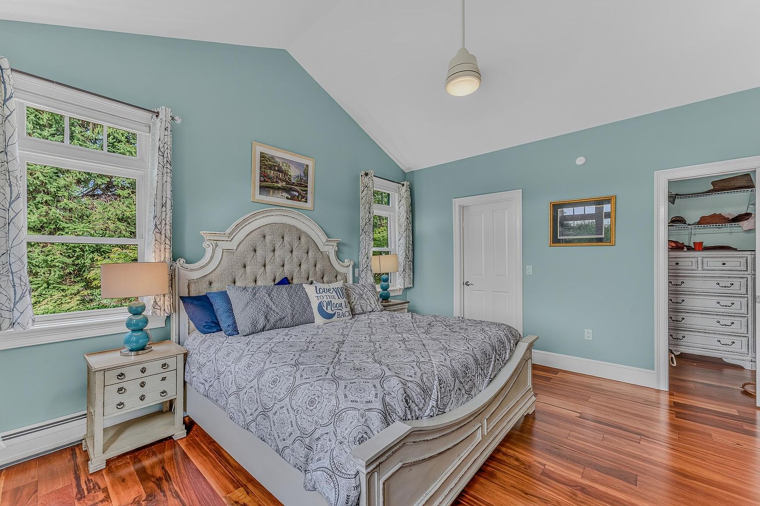 This primary bedroom features a vaulted ceiling and light blue walls, creating a serene and airy atmosphere. The room is anchored by a large, tufted upholstered headboard and a patterned bedspread, flanked by two matching light-toned nightstands with blue glass lamps. A doorway leads into a walk-in closet, while rich-toned wood flooring runs throughout the space, providing a warm contrast to the cool wall color.