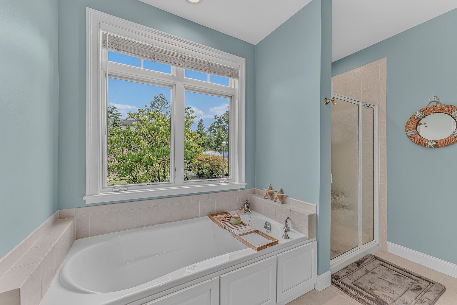 This bright and airy bathroom features a large soaking tub positioned beneath a wide window that offers a view of the outdoors. The space is finished with light blue walls, neutral tile surrounds, and a separate glass-enclosed shower stall. A decorative round mirror with a rope-style frame adds a coastal touch to the serene, well-lit room.