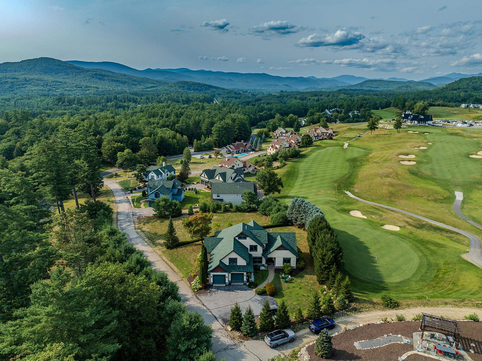 This high-angle aerial view captures a residential property nestled alongside a lush green golf course, set against a backdrop of rolling mountains and a clear blue sky. The home features a multi-gabled roofline, a paved driveway, and a well-maintained lawn, with neighboring houses visible along the winding road. The perspective emphasizes the scenic, community-oriented location and the expansive natural surroundings of the property.