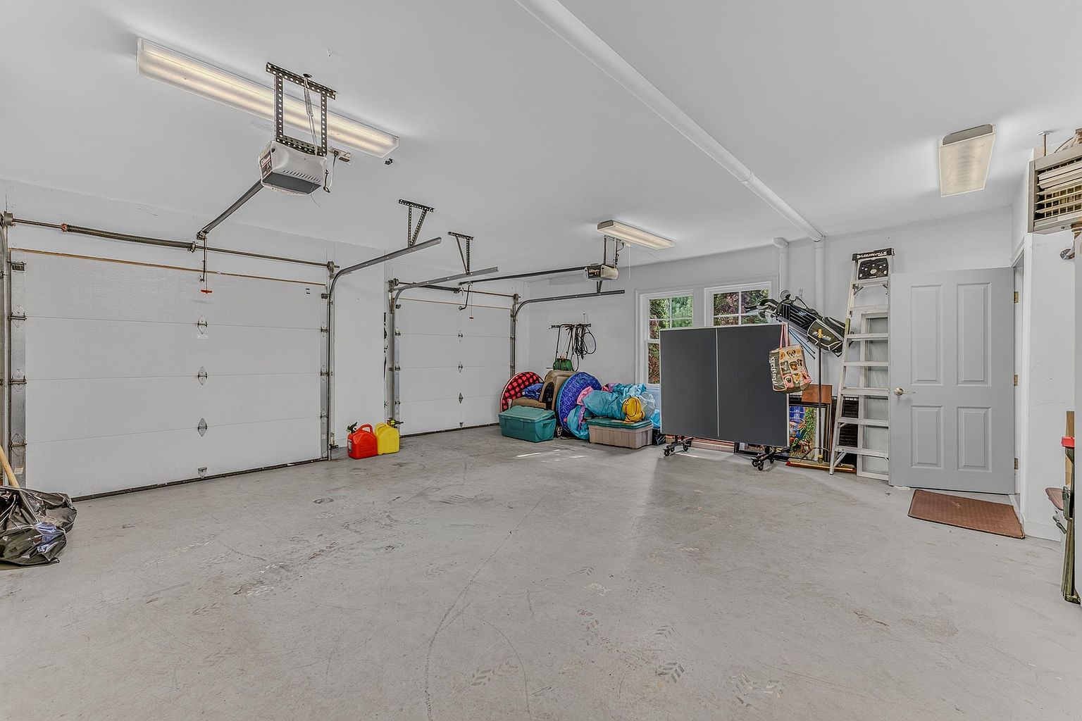 This spacious two-car garage features a clean, neutral aesthetic with white walls and a light-colored concrete floor. The area is equipped with overhead lighting, two garage door openers, and a side entry door, providing ample room for storage or recreational activities like the ping-pong table currently set up. The perspective is wide-angle, capturing the full depth and functional layout of the garage space.