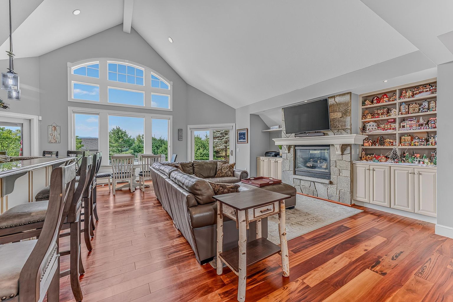 This open-concept living room features rich wood flooring and a vaulted ceiling that enhances the sense of space. A stone-clad chimney breast serves as the focal point, housing a fireplace and a television above, while built-in shelving displays a collection of decorative items. Large windows and glass doors provide abundant natural light and views of the outdoors, creating a bright and inviting atmosphere.