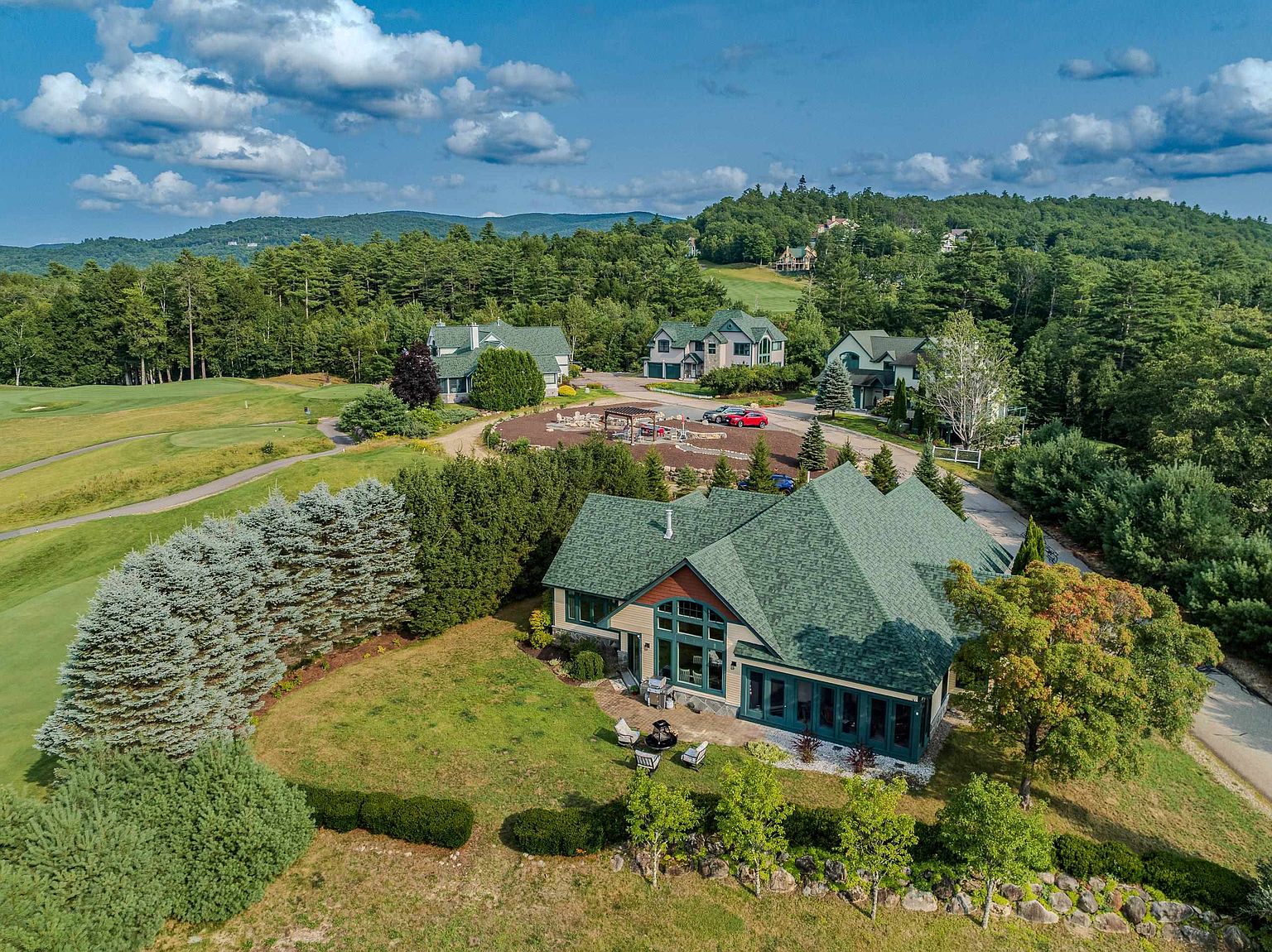 This high-angle aerial view captures a sprawling, multi-gabled residence nestled within a lush, green landscape adjacent to a golf course. The home features a dark green shingled roof, a stone and siding exterior, and a rear patio area equipped with outdoor seating. The surrounding property includes manicured lawns, mature trees, and a winding driveway that connects to neighboring homes in a quiet, scenic community.