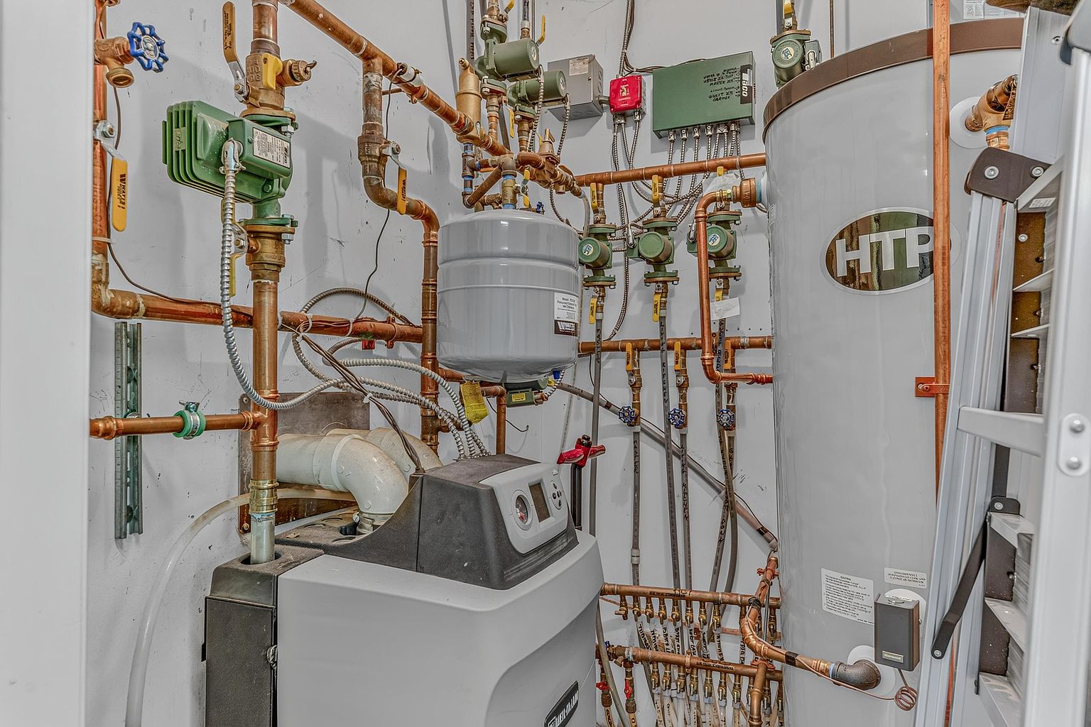 This image displays a complex mechanical room featuring a high-efficiency boiler system, a large HTP water storage tank, and an expansion tank. The setup includes intricate copper piping, multiple green circulation pumps, and various control valves, showcasing a professional-grade residential heating and plumbing installation. The perspective is a direct, eye-level view of the utility equipment, emphasizing the technical infrastructure of the home.
