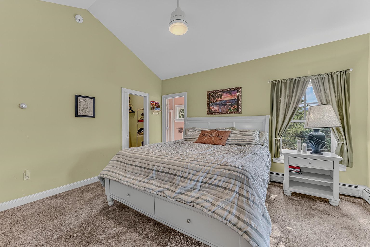 This primary bedroom features a vaulted ceiling and neutral-toned walls, creating a bright and airy atmosphere. A white bed frame with a striped comforter serves as the focal point, complemented by a matching white nightstand and a table lamp. The room includes a window with draped curtains and provides access to an adjacent closet and bathroom area.