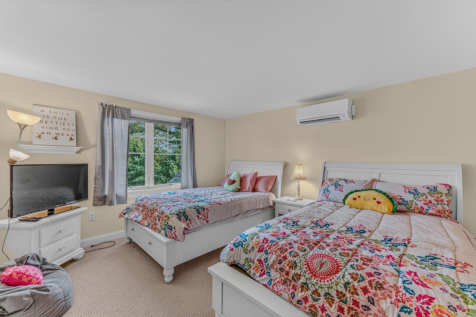This bright and cheerful bedroom features two twin beds with colorful, floral-patterned bedding and whimsical plush pillows. The room is furnished with a white dresser supporting a television, a matching nightstand with a lamp, and a floor lamp, all set against neutral-toned walls. A window with grey curtains allows natural light to fill the space, which is also equipped with a wall-mounted air conditioning unit.