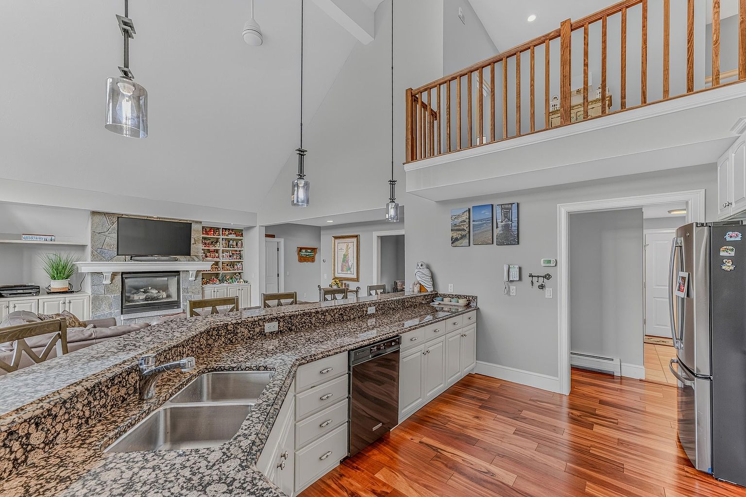 This open-concept kitchen features a large, multi-level granite island with an integrated double sink and dark-toned cabinetry. The space flows into a living area highlighted by a stone-clad chimney breast and a vaulted ceiling, while a wooden staircase railing overlooks the kitchen from an upper level. Warm wood flooring runs throughout the area, creating a cohesive and inviting atmosphere.
