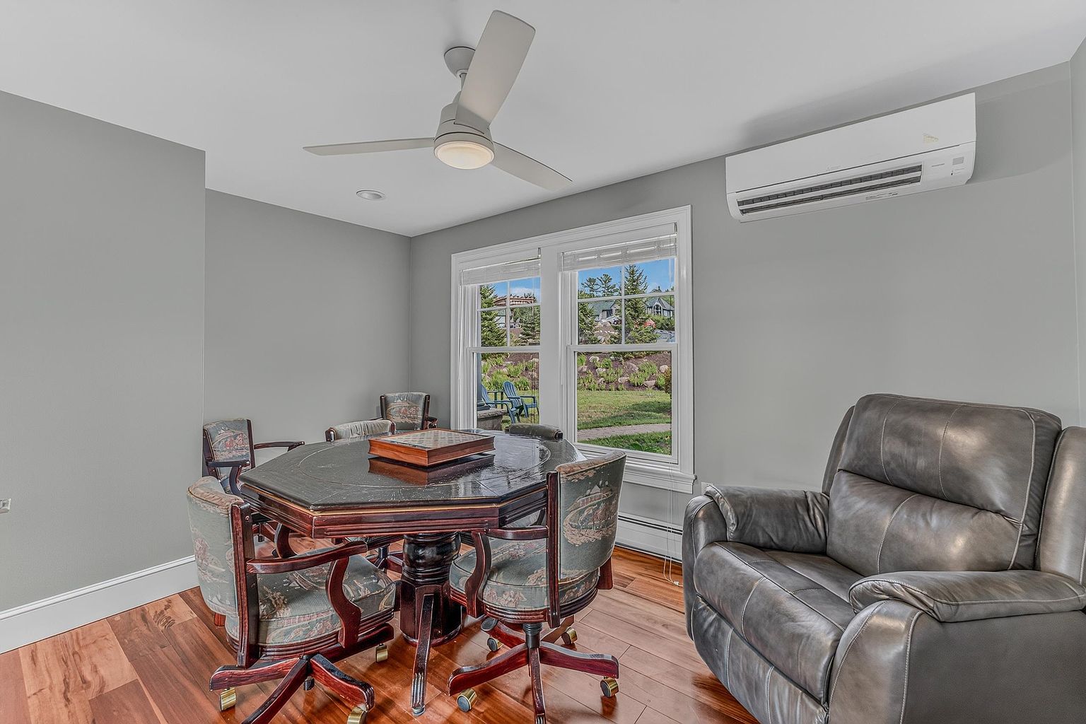 This room features a dark wood game table surrounded by four upholstered chairs, positioned near a pair of windows that look out onto a landscaped yard. A large, comfortable leather recliner sits to the right, while a ceiling fan and a wall-mounted air conditioning unit provide climate control. The space is finished with warm-toned wood flooring and neutral gray walls, creating a cozy and functional area for leisure or work.