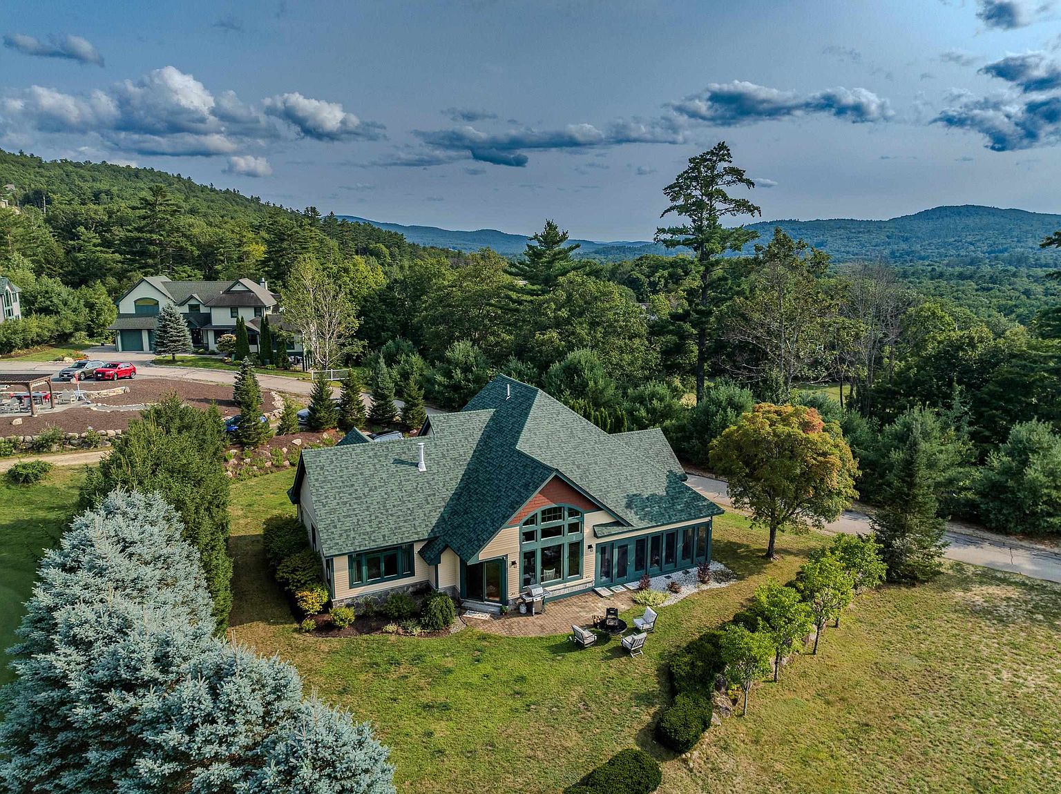 This aerial perspective captures a charming, multi-gabled home nestled in a lush, green landscape with rolling mountains in the distance. The house features a prominent green shingled roof, a stone-accented facade, and a large central window arrangement that floods the interior with natural light. A paved patio area with outdoor seating sits at the rear, surrounded by manicured lawns and mature trees, creating a serene and inviting atmosphere.