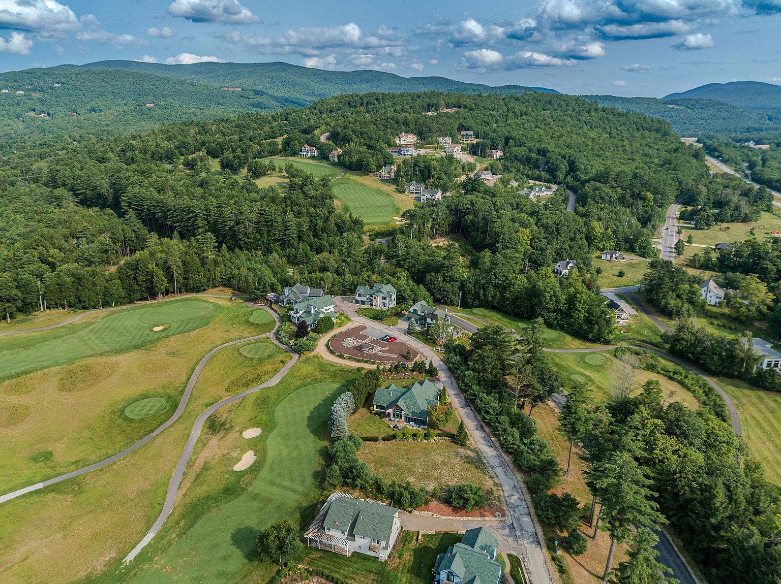 This high-angle aerial perspective captures a sprawling residential community nestled within a lush, green landscape adjacent to a golf course. The scene features several multi-story homes with dark green roofs, winding paved roads, and manicured fairways that blend seamlessly into the surrounding dense forest and rolling mountains. The composition provides a comprehensive view of the property's integration with its natural environment, emphasizing the quiet, scenic setting.