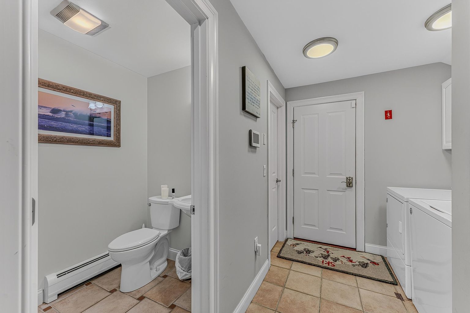 This functional space serves as a combined laundry room and powder room, featuring light-colored walls and neutral-toned tile flooring. To the left, a small bathroom contains a white toilet and a pedestal sink, while the right side houses a white washer and dryer set. A white door leads to an exterior entrance, accented by a ski-themed rug, creating a practical and utilitarian transition area.