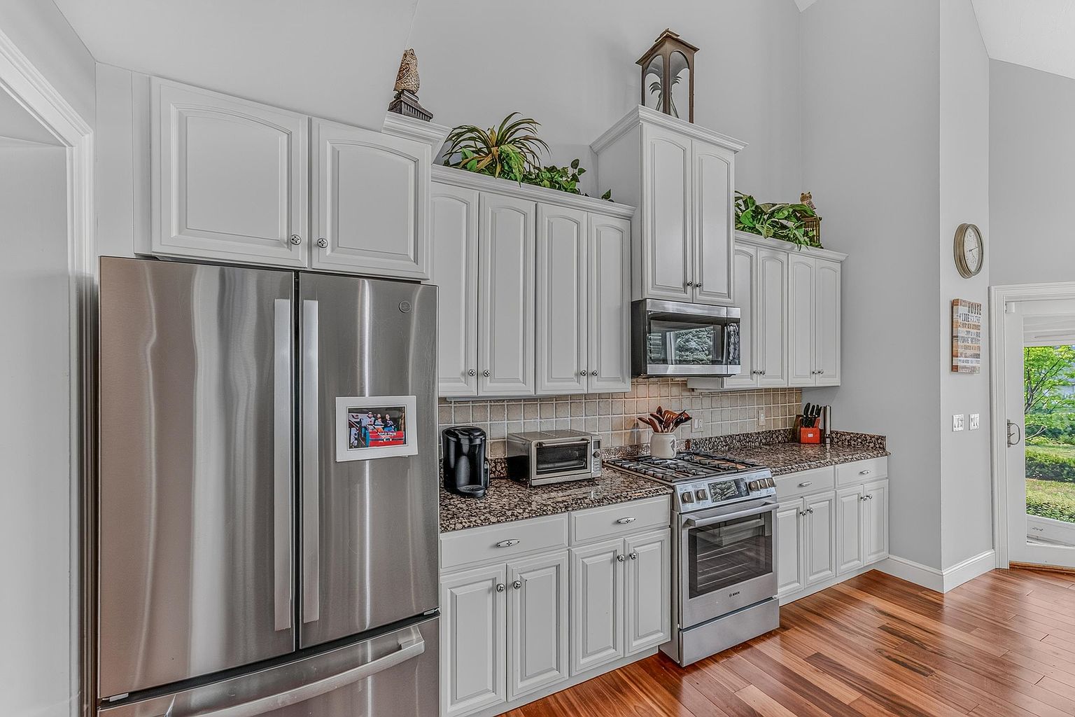 This bright kitchen features white cabinetry paired with speckled stone countertops and a neutral tiled backsplash. The space is equipped with stainless steel appliances, including a large refrigerator and a gas range, and is accented by warm wood flooring. A tall, vaulted ceiling adds a sense of openness, while decorative plants and a wall clock provide a lived-in, welcoming atmosphere.
