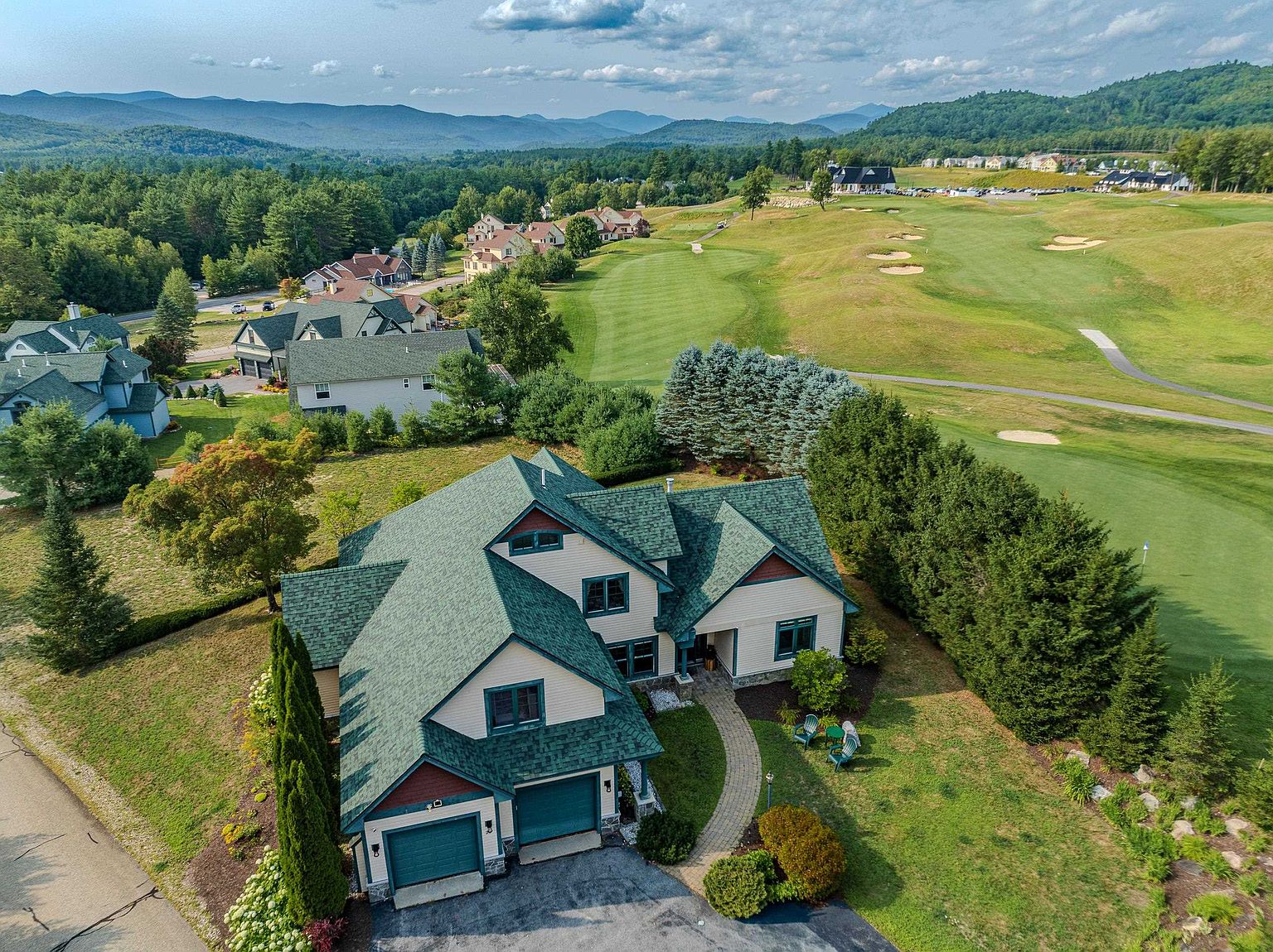 This high-angle aerial view captures a multi-level suburban home with a distinctive green shingled roof and light-colored siding, situated directly adjacent to a lush golf course. The property features a paved driveway leading to a two-car garage, a stone-paved walkway to the front entrance, and a well-maintained lawn bordered by mature trees. In the background, rolling mountains and a clear blue sky provide a scenic, expansive backdrop for this residential setting.