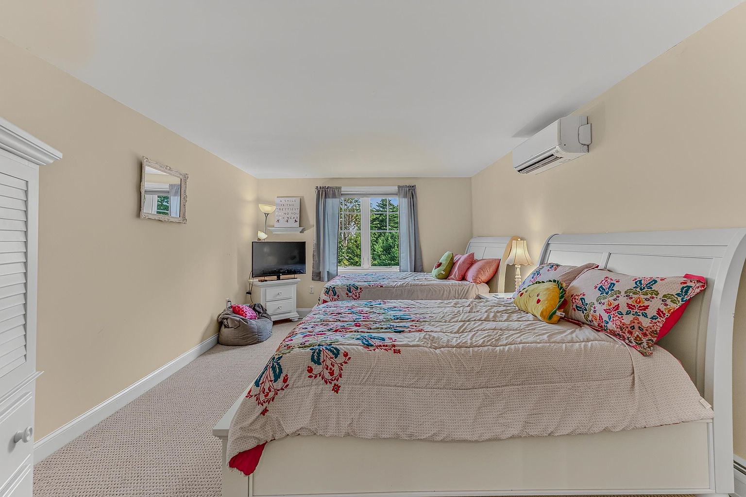 This spacious bedroom features two beds with white frames and patterned bedding, creating a comfortable and inviting atmosphere. The room is illuminated by natural light from a central window and includes a small television stand, a beanbag chair, and a wall-mounted air conditioning unit. The neutral wall color and carpeted flooring provide a clean, versatile backdrop for the space.