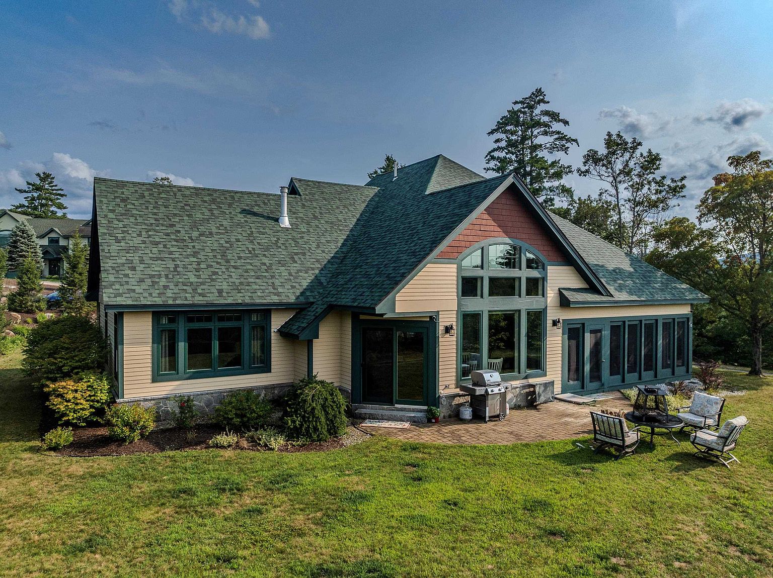 This elevated perspective captures the rear exterior of a multi-level home featuring a combination of light-toned siding and dark green trim. The house showcases a prominent gabled roofline with green shingles, a large arched window assembly, and a stone-accented foundation. A paved patio area extends from the home, furnished with outdoor seating and a grill, overlooking a spacious, well-maintained lawn.
