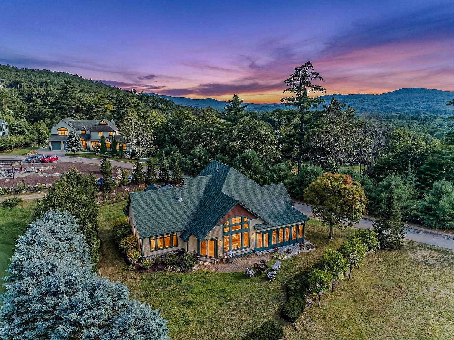 This high-angle aerial shot captures a sprawling, multi-gabled home nestled in a lush, mountainous landscape during a vibrant sunset. The property features a stone-paved patio area with outdoor seating, surrounded by manicured lawns and mature trees, while a secondary residence is visible in the background. The dramatic purple and orange sky provides a picturesque backdrop for this serene, private estate.