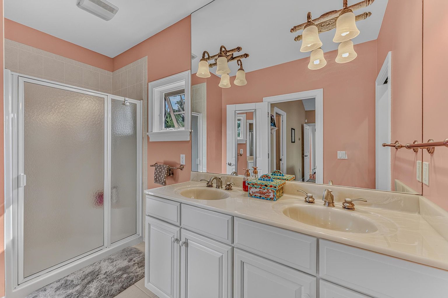 This bathroom features a dual-sink vanity with a light-toned countertop and white cabinetry, set against soft peach-colored walls. A large, wall-to-wall mirror reflects the space, including a glass-enclosed shower stall with textured privacy glass and a small window above it. The room is illuminated by two decorative light fixtures with multiple glass shades, creating a bright and traditional aesthetic.