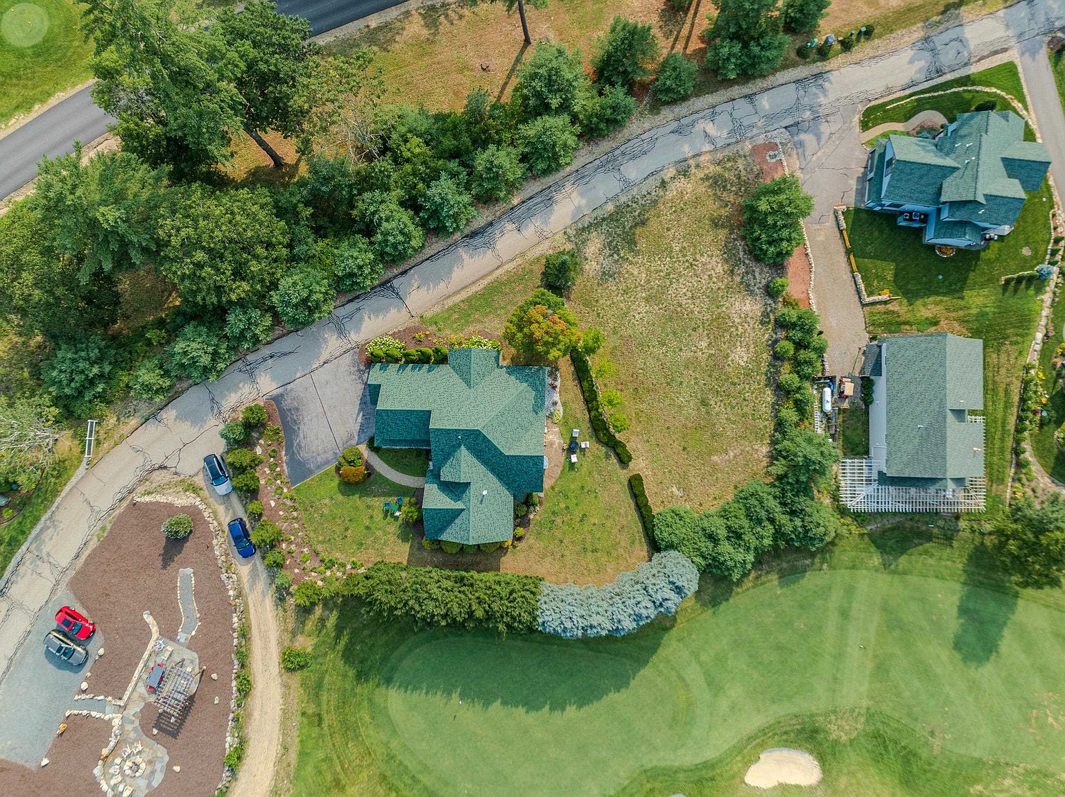 This high-angle aerial perspective captures a residential property nestled alongside a golf course, showcasing a sprawling green-roofed home with an attached driveway. The property features a well-manicured lawn, mature landscaping, and a stone-paved outdoor fire pit area, all bordered by lush trees and a winding road. The layout highlights the home's integration into the surrounding landscape, emphasizing its proximity to the golf course green and sand trap.