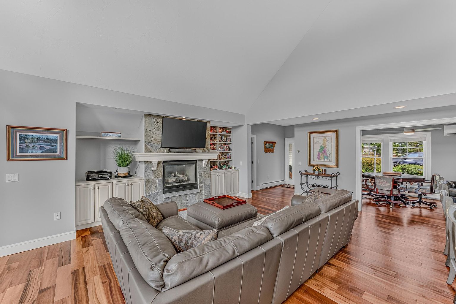 This spacious living room features a vaulted ceiling and warm-toned wood flooring, centered around a stone-clad chimney breast with a built-in fireplace. A large, neutral-toned sectional sofa and matching ottoman provide comfortable seating, while built-in cabinetry and shelving flank the fireplace. The open-concept layout flows seamlessly into an adjacent dining area, creating an inviting and cohesive living space.