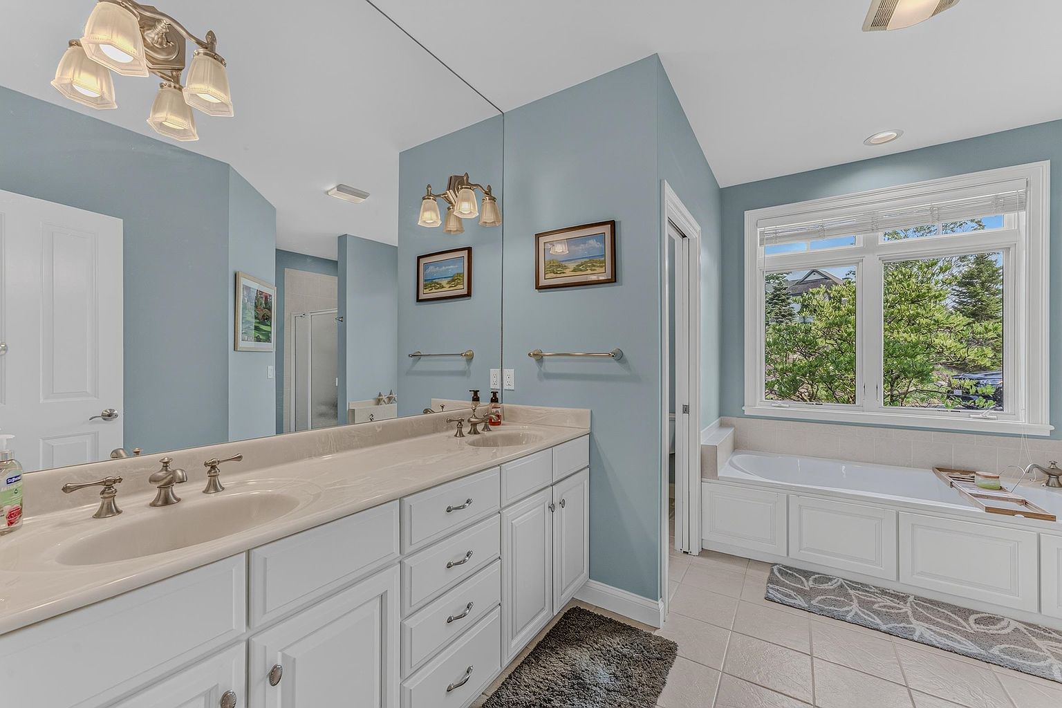 This primary bathroom features a dual-sink vanity with white cabinetry and a light-toned countertop, complemented by a large wall-to-wall mirror. To the right, a deep soaking tub is positioned beneath a large window that allows for ample natural light. The room is finished with light blue walls and neutral floor tiles, creating a bright and serene atmosphere.