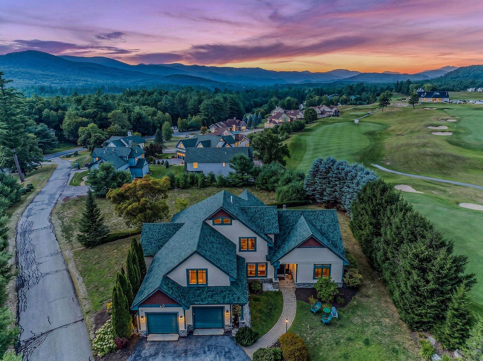 This stunning aerial perspective captures a multi-story suburban home nestled within a scenic golf course community at sunset. The residence features a complex roofline with dark shingles, a two-car garage, and a stone-paved walkway leading to the front entrance, all surrounded by lush green landscaping and mature trees. In the background, rolling mountains are silhouetted against a vibrant, multi-colored twilight sky, creating a serene and picturesque atmosphere.