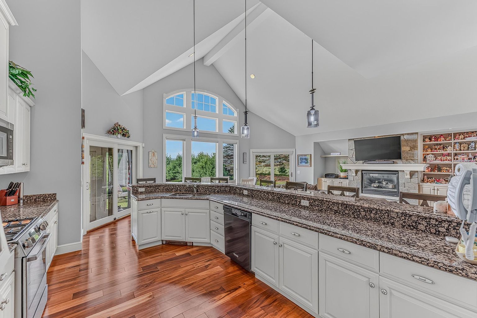 This spacious, open-concept kitchen features white cabinetry paired with dark, patterned stone countertops and warm-toned hardwood flooring. The room is highlighted by a vaulted ceiling and a large window arrangement that allows for abundant natural light, while a stone-clad chimney breast with a fireplace adds a cozy focal point to the adjacent living area. The perspective captures the expansive layout, showcasing the seamless transition between the kitchen workspace and the comfortable living space.