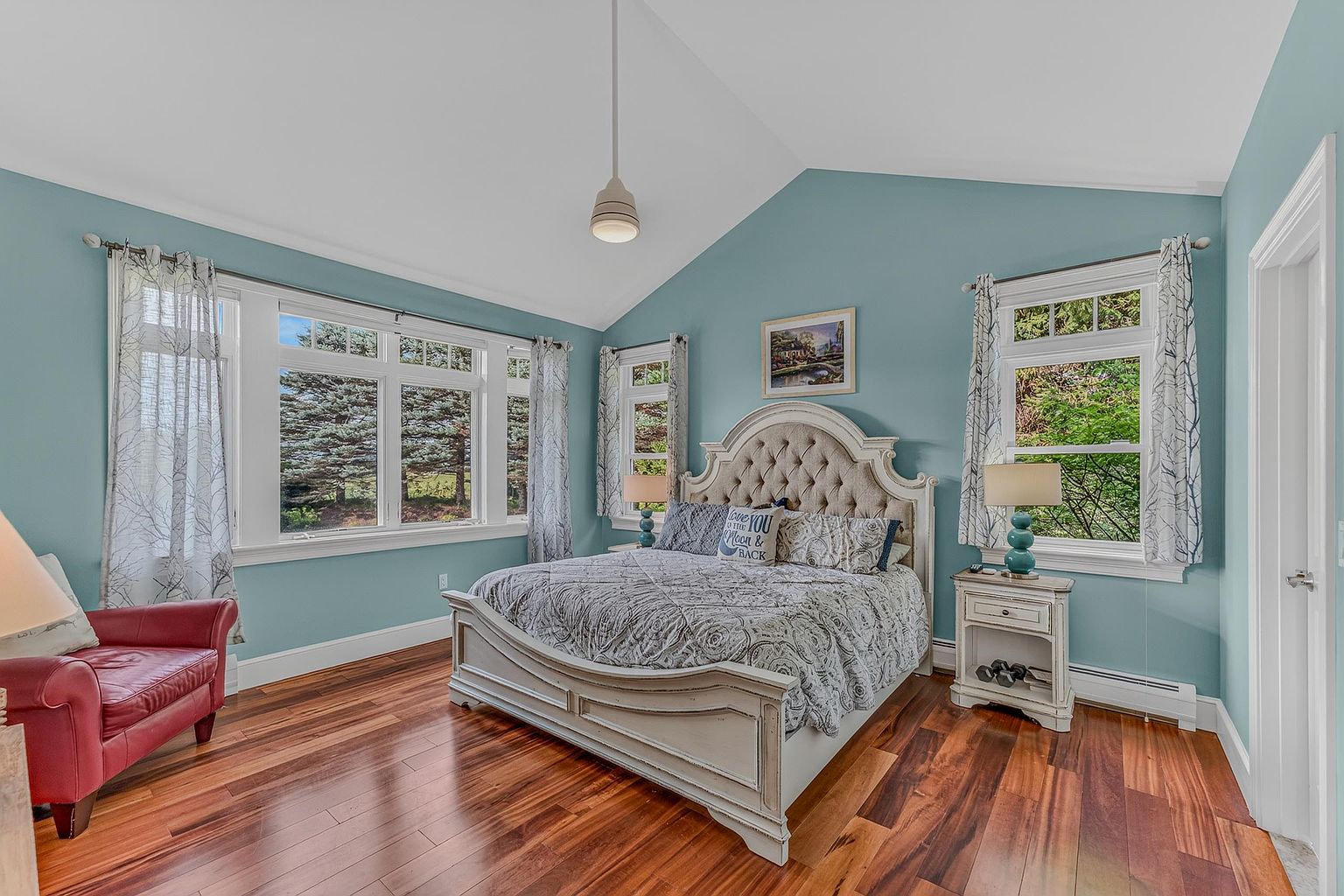 This primary bedroom features a vaulted ceiling and light blue walls, creating a serene and airy atmosphere. The room is anchored by a tufted, arched headboard bed with patterned bedding, flanked by two matching nightstands with lamps. A red leather armchair sits in the corner, and the space is illuminated by a central pendant light and natural light streaming through multiple windows.