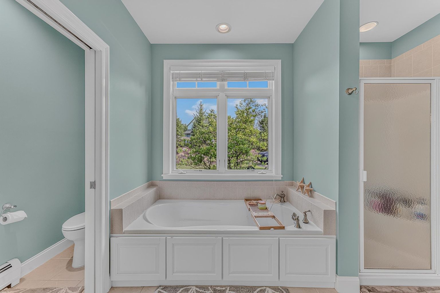 This bright and airy bathroom features a large soaking tub centered beneath a window that offers a view of the outdoors. The space is finished with light blue walls, white cabinetry, and neutral tile surrounds, creating a clean and tranquil atmosphere. A glass-enclosed shower is visible to the right, while a toilet is tucked into the corner on the left.