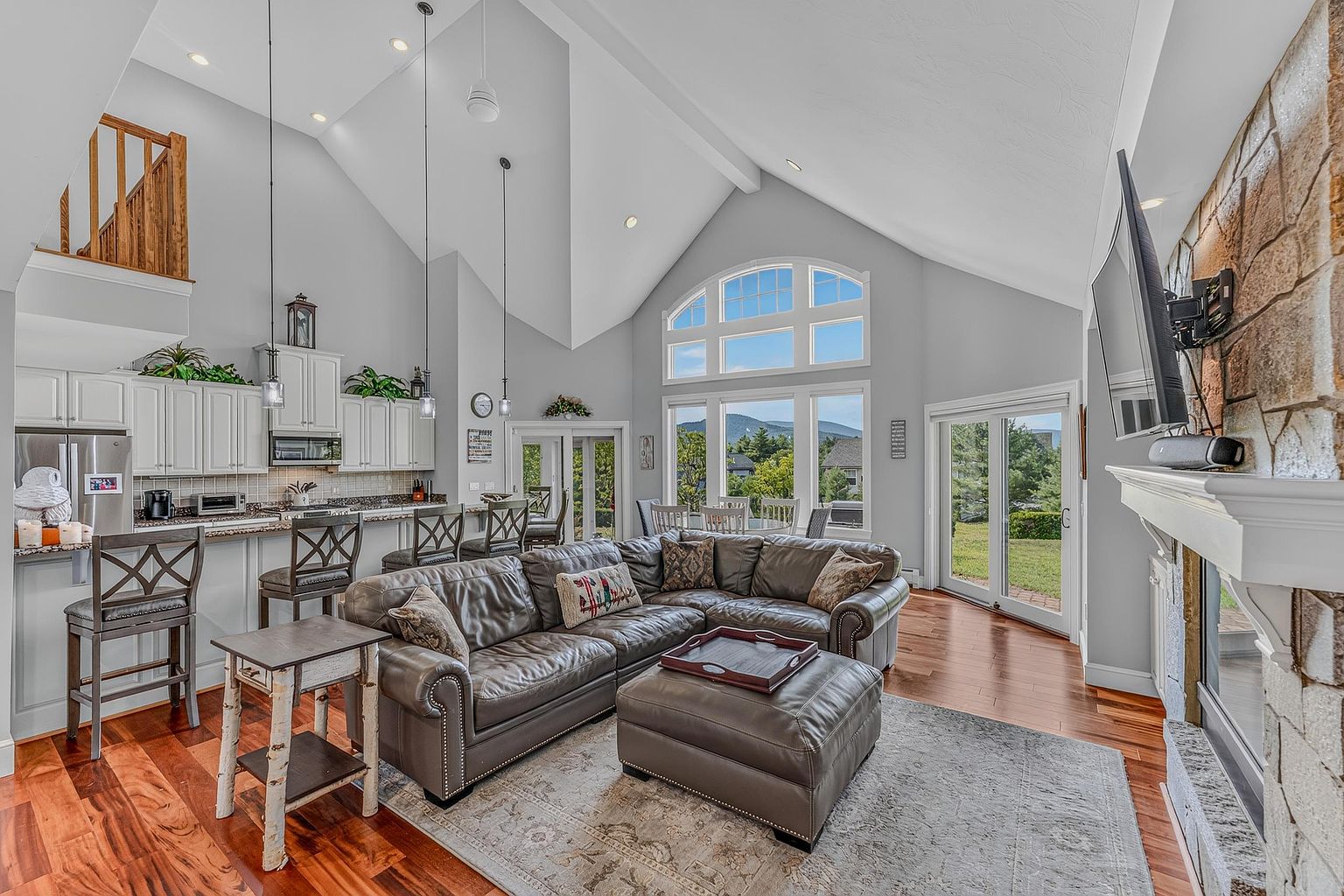 This open-concept living space features a vaulted ceiling and a large sectional sofa centered around a stone-clad chimney breast with a mounted television. The room flows seamlessly into a kitchen area with a breakfast bar and stools, while a wall of windows provides abundant natural light and views of the outdoors. The warm wood flooring and neutral color palette create an inviting and spacious atmosphere suitable for entertaining.