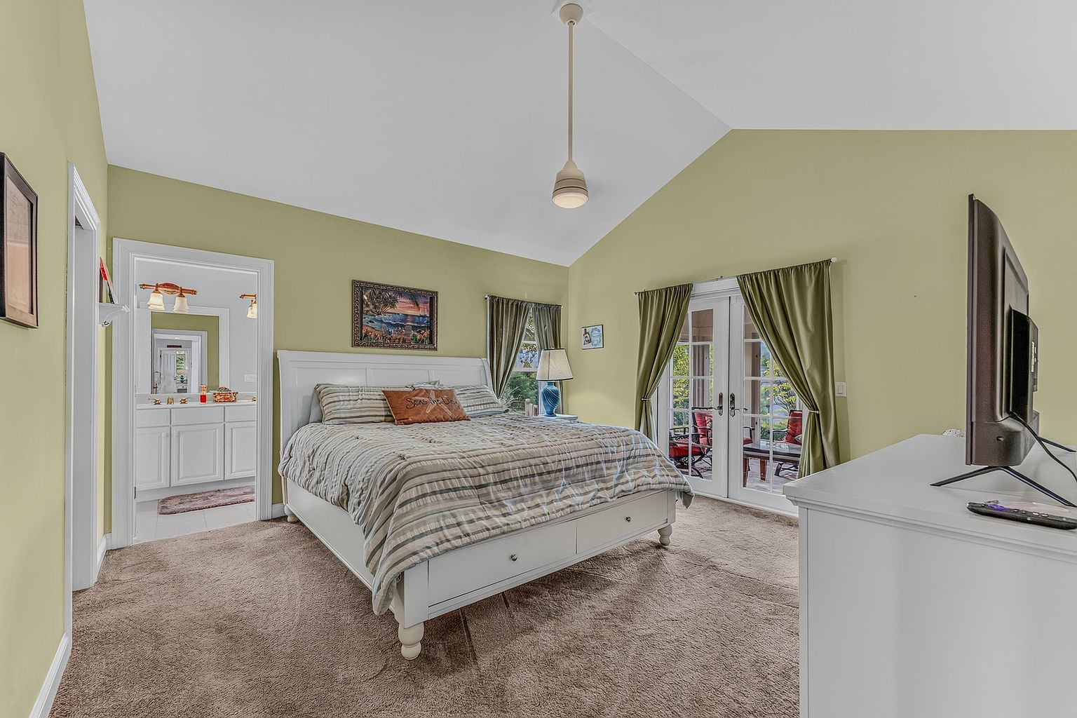 This spacious primary bedroom features a vaulted ceiling and neutral-toned walls, creating an airy and inviting atmosphere. A large bed with a white frame serves as the focal point, complemented by a dresser with a television and a doorway leading to an en-suite bathroom. French doors with green curtains open to a private outdoor space, allowing natural light to fill the room.
