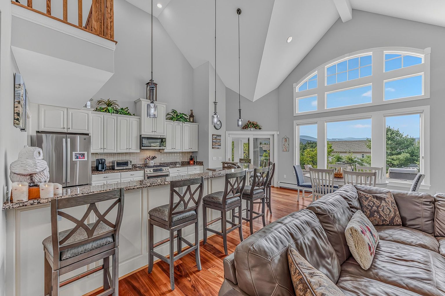 This open-concept kitchen features white cabinetry, granite countertops, and a breakfast bar with four stools, seamlessly flowing into a living area. The space is defined by its vaulted ceilings, tall windows that provide abundant natural light, and warm wood flooring. The perspective captures the kitchen's functional layout and its connection to the adjacent dining and living spaces, creating an inviting and airy atmosphere.