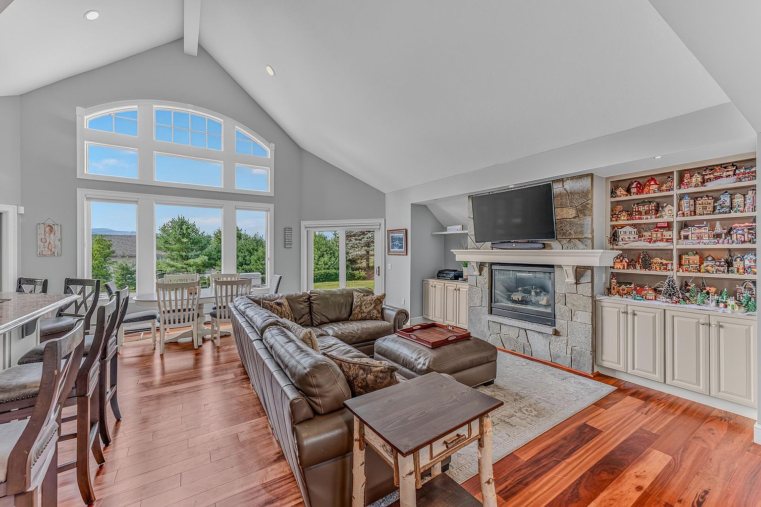 This spacious living room features a vaulted ceiling and warm wood flooring, creating an inviting and open atmosphere. A large stone-clad chimney breast serves as the focal point, housing a fireplace and a mounted television, while built-in shelving displays a collection of decorative village houses. Large windows and a glass door provide ample natural light and views of the surrounding greenery, seamlessly connecting the indoor living space with the outdoors.