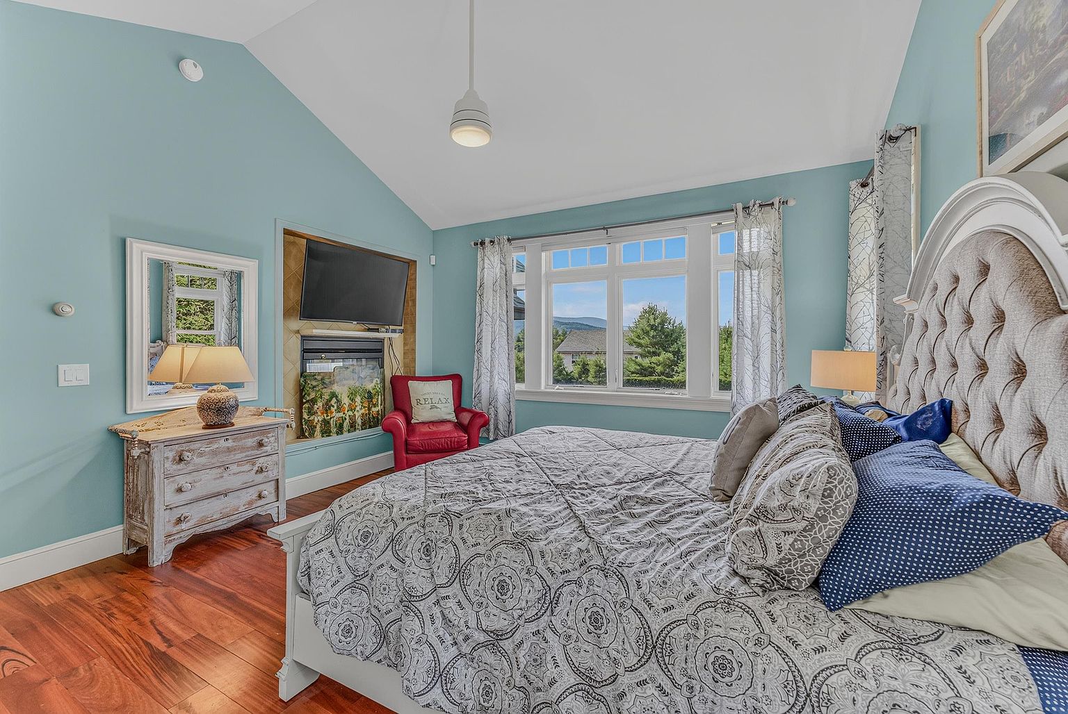 This primary bedroom features a vaulted ceiling and light blue walls, creating a bright and airy atmosphere. A large bed with a tufted headboard serves as the focal point, while a red armchair sits in the corner near a recessed area containing a television and a fireplace. Large windows provide natural light and a view of the surrounding landscape, complemented by warm wood flooring.